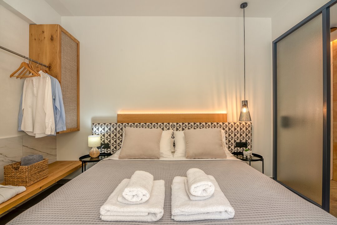 Cozy bedroom with king bed, geometric headboard, rattan lamp, open wardrobe, and frosted glass bathroom partition at Nusa Balos, Crete